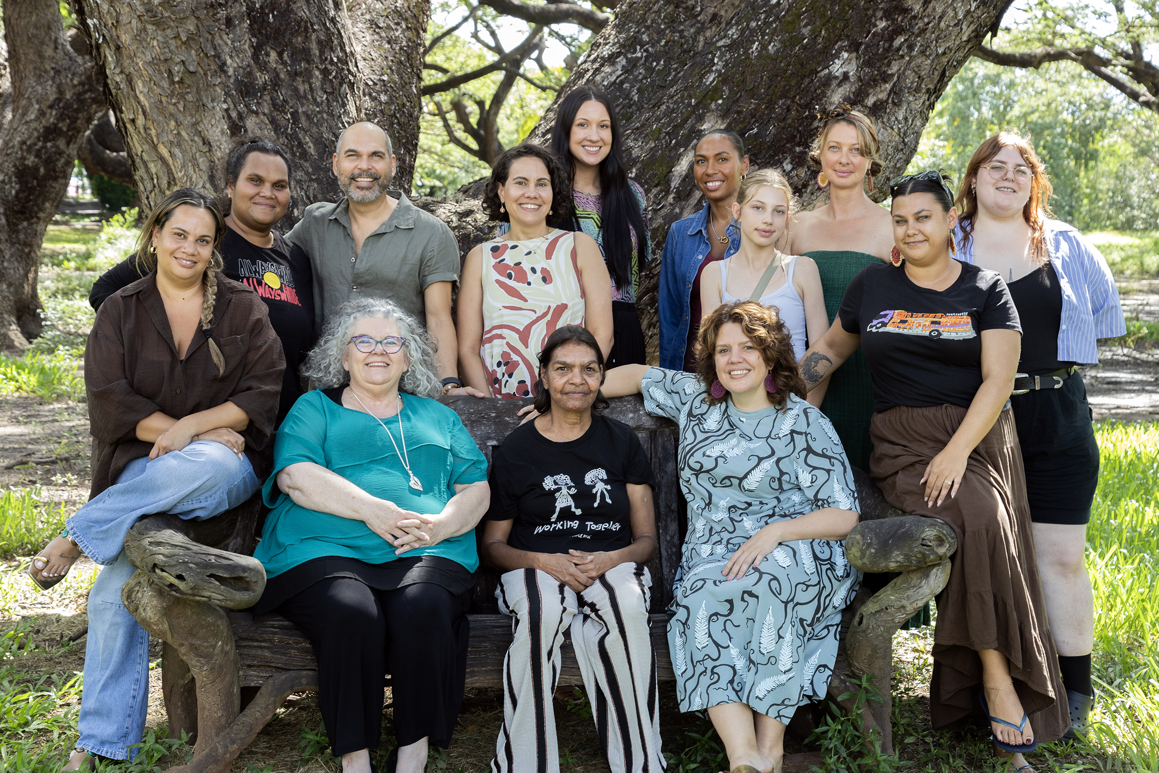 Group of people gathered outdoors beneath large tree, posing together around wooden bench.